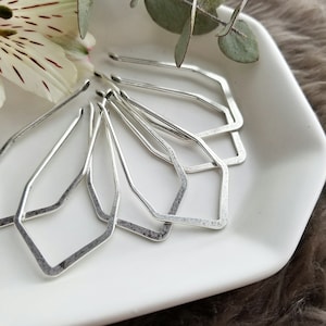 May include: A collection of geometric silver earrings in various shapes, including diamond and hexagonal designs. The earrings have a hammered texture and are arranged on a white square plate. A white flower with red markings and green leaves are in the background.