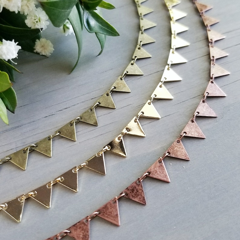May include: Three gold and copper triangle chain necklaces. The chains are layered on a light gray surface.