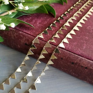 Pode incluir: Três fios de corrente dourada com elos em forma de triângulo. A corrente está sobre um livro bordô com folhas verdes.