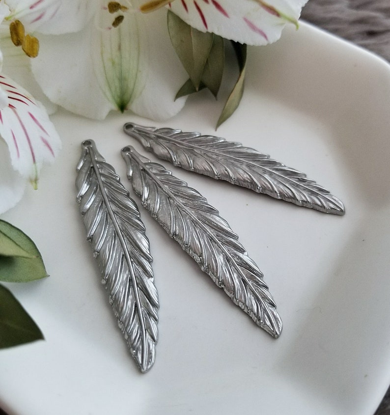 Op de afbeelding: Drie zilveren metalen veerbedels met een gedetailleerd, gestructureerd ontwerp. De bedels liggen op een wit oppervlak.