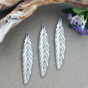 Op de afbeelding: Drie zilveren veerbedels met een gedetailleerd ontwerp. De veren zijn ongeveer 5 cm lang en hebben een gat aan de bovenkant om aan sieraden te bevestigen.