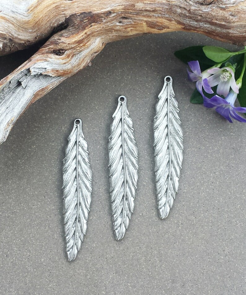 Op de afbeelding: Drie zilveren veerbedels met een gedetailleerd ontwerp. De veren zijn ongeveer 5 cm lang en hebben een gat aan de bovenkant om aan sieraden te bevestigen.