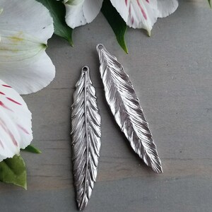 Op de afbeelding: Twee zilveren verencharms met een gedetailleerd, gestructureerd ontwerp. De charms hebben een lus aan de bovenkant om aan sieraden of andere knutselwerken te bevestigen.