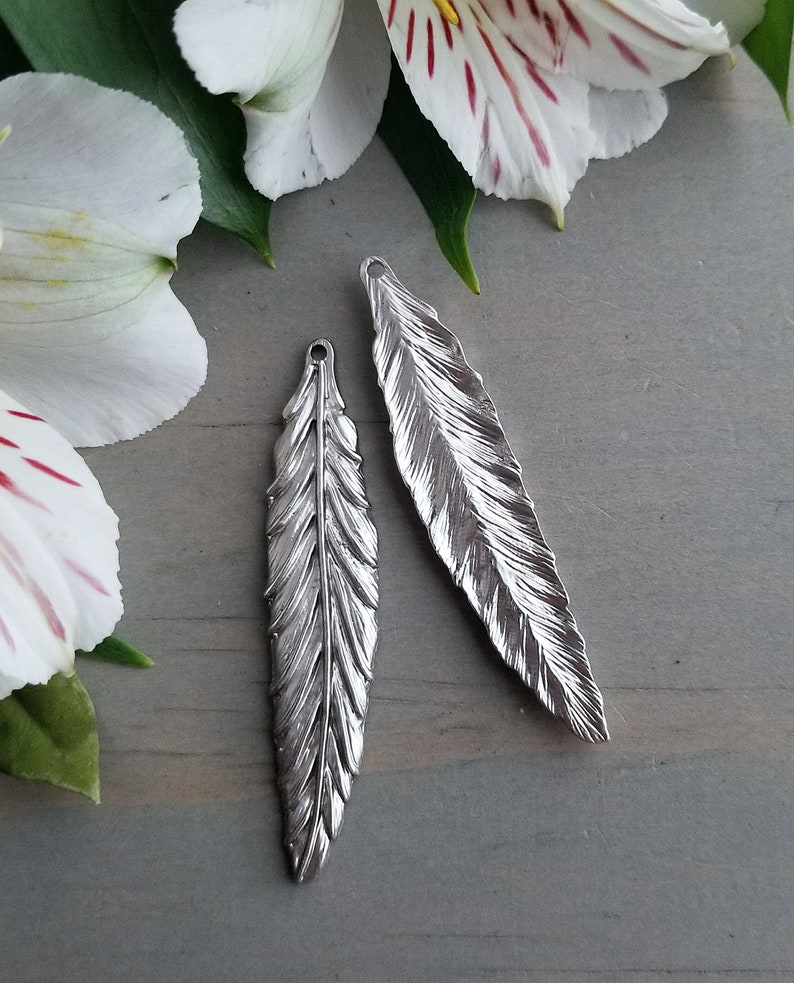 Op de afbeelding: Twee zilveren verencharms met een gedetailleerd, gestructureerd ontwerp. De charms hebben een lus aan de bovenkant om aan sieraden of andere knutselwerken te bevestigen.