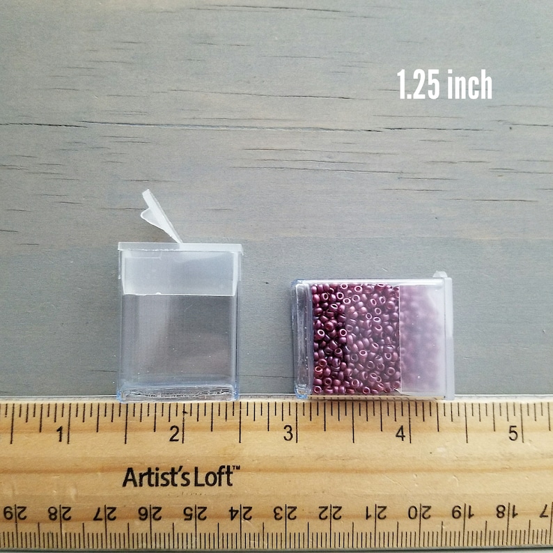 May include: Two clear plastic containers with lids, one open and one closed, filled with small, dark red beads. The containers are 1.25 inches wide and are measured on a wooden ruler.