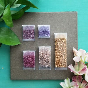 May include: Six small clear containers filled with various colors of seed beads. The beads are in shades of purple, gold, pink, and white. The containers are arranged in two rows of three on a gray surface.