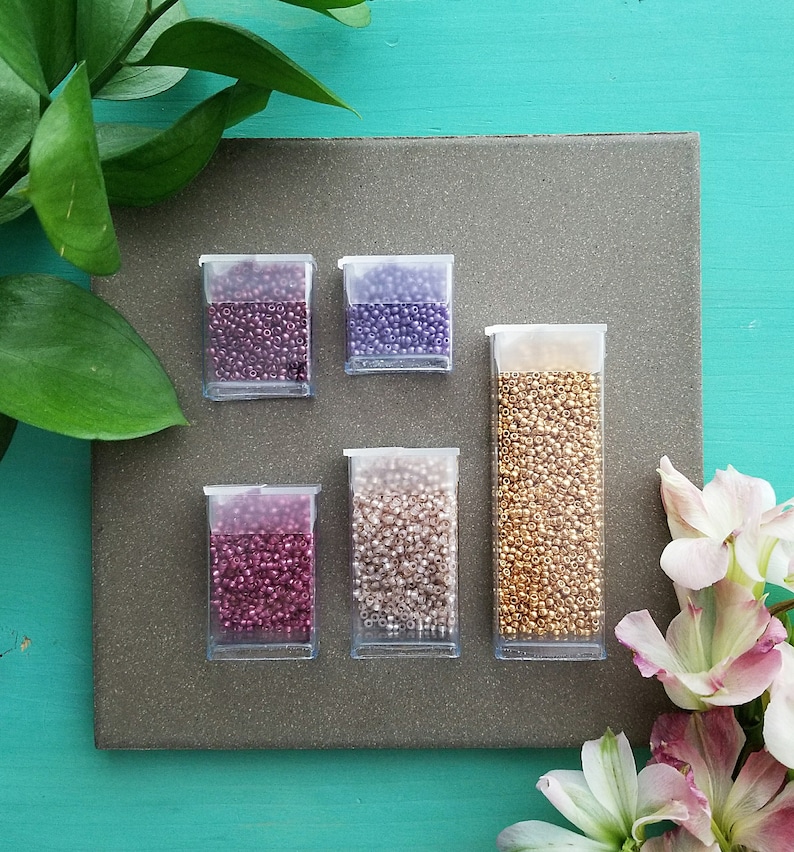 May include: Six small clear containers filled with various colors of seed beads. The beads are in shades of purple, gold, pink, and white. The containers are arranged in two rows of three on a gray surface.