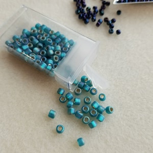 May include: A clear plastic container filled with teal blue seed beads. Some beads have spilled out of the container onto a white surface.