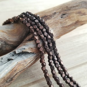 May include: A strand of dark brown metallic beads with a faceted, hexagonal shape. The beads are strung on a wire and are arranged in a loose coil.