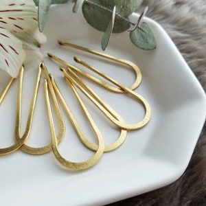 May include: A set of six gold-toned hair clips in a teardrop shape. The clips are arranged in a fan shape on a white surface.