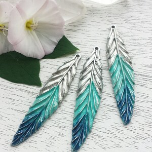 May include: Three silver feather charms with a gradient of blue and teal paint. The feathers are arranged in a row on a white wooden surface.