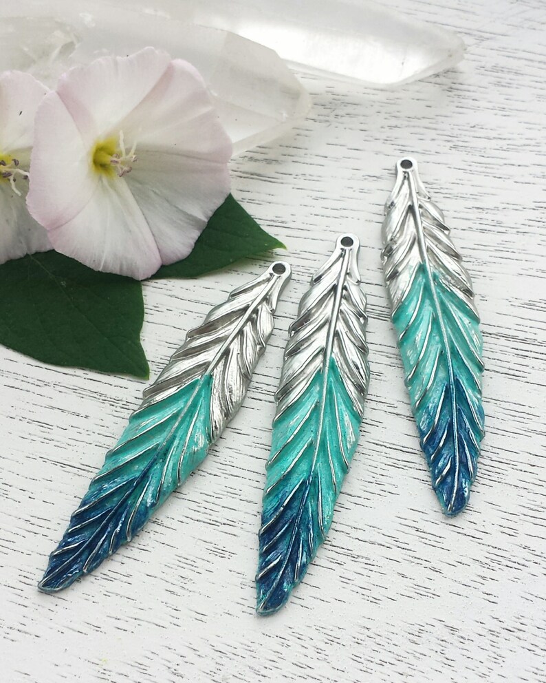 May include: Three silver feather charms with a gradient of blue and teal paint. The feathers are arranged in a row on a white wooden surface.