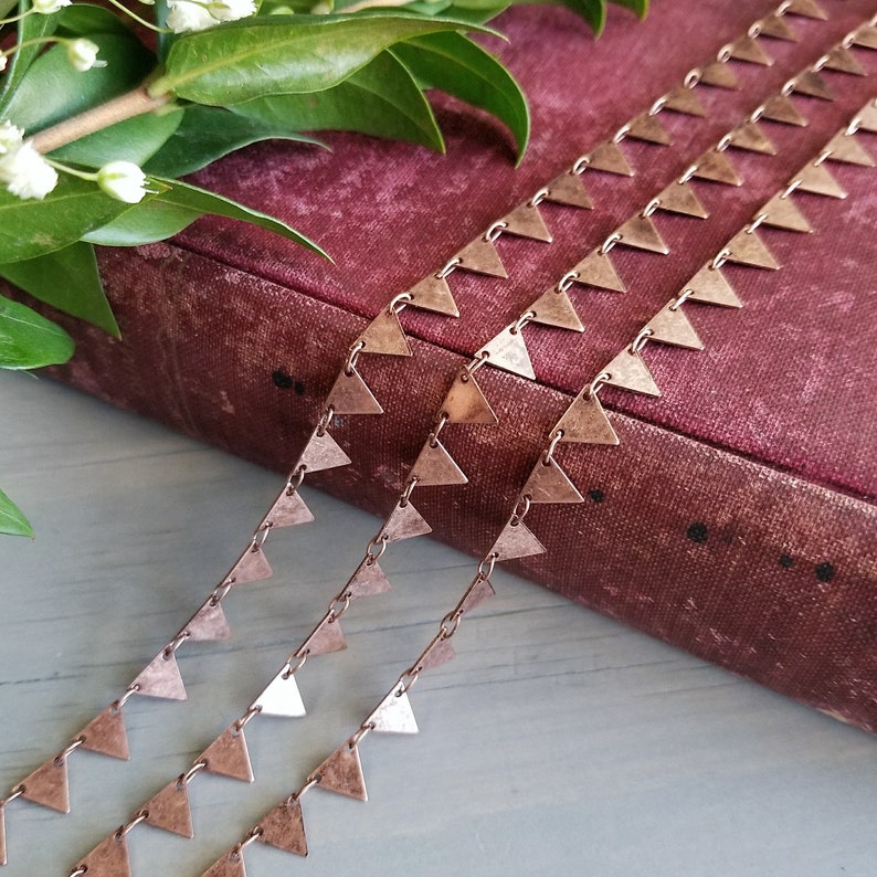 May include: Three strands of copper chain with triangle-shaped charms. The chain is lying on a burgundy book.