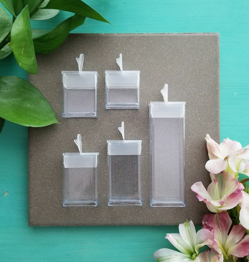 May include: Five clear plastic boxes with hinged lids. The boxes are different sizes and are arranged in a row on a gray surface.