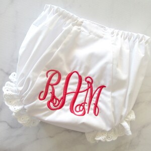 Op de afbeelding: Witte babybloomers met een roze geborduurde monogram "RAM" en witte kant.