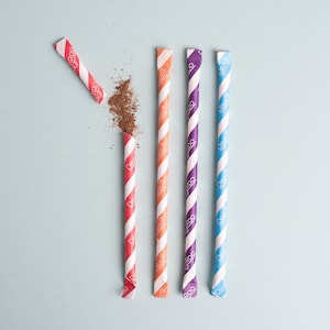 May include: Five colorful paper straws with red, orange, purple, and blue stripes. The straws are arranged vertically on a light blue background. Some of the straws have the word "Dezi" printed on them.