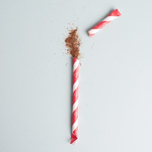 May include: A red and white striped paper straw with a red and white striped paper straw on a light blue background. The straw is broken in half and the top half is laying on the bottom half. There is a pile of brown powder on the bottom half of the straw.