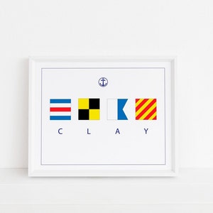 May include: A white framed print with the word "CLAY" spelled out using nautical signal flags. The flags are red and yellow striped, black and yellow checkered, blue and white, and red and white striped. An anchor symbol is above the flags.