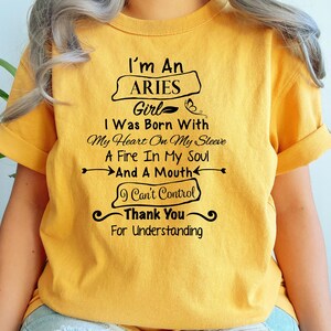 Könnte beinhalten: Senfgelbes T-Shirt mit schwarzem Text. Der Text lautet "I'm An Aries Girl", gefolgt von einem Zitat über die Geburt mit einem Feuer in der Seele und einem unkontrollierbaren Mund. Das Shirt hat kurze Ärmel.