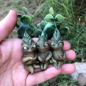 Könnte beinhalten: Drei kleine grüne und braune Tonfiguren mit blattartigen Merkmalen und großen Augen. Die Figuren sitzen auf einer Hand, mit grünen, blättrigen Stielen dahinter.