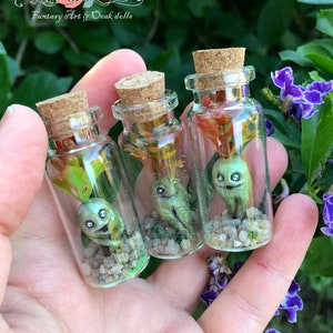 Puede incluir: Tres pequeñas botellas de vidrio con tapones de corcho, cada una contiene una figura humanoide verde con una cara y una pequeña planta. Las botellas están llenas de pequeñas piedras y purpurina.