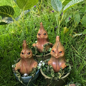 Könnte beinhalten: Drei kleine, braune, humanoide Figuren mit großen Augen und langen, dünnen Hälsen sind in Terrakotta-Töpfen eingetopft. Die Figuren sitzen aufrecht mit gekreuzten Armen. Die Töpfe sind von grünem Gras und Blättern umgeben.