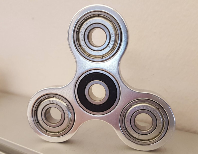 May include: A silver fidget spinner with three arms and three ball bearings. The ball bearings have the text "BC Precision 608ZZ" engraved on them.