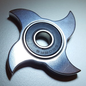 Pode incluir: Um spinner fidget de metal prateado com três pontas e um rolamento preto com a palavra "SOARS" impressa nele.