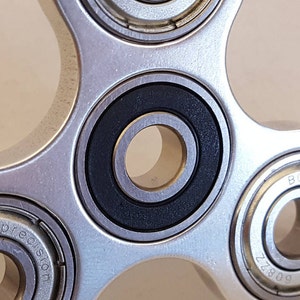 May include: A silver fidget spinner with five arms and five bearings. The bearings are black and silver and have a smooth, polished finish.