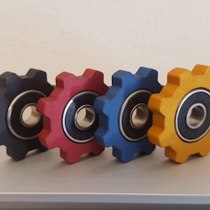 May include: Four fidget spinners in different colors: black, red, blue, and gold. Each spinner has a black bearing in the center and a gear-like design.