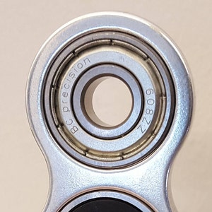 May include: Close-up of a silver fidget spinner with a bearing that reads "BC Precision ZZ 809".