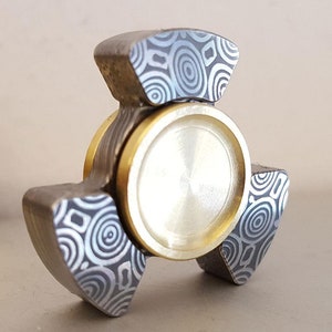 May include: A three-lobed fidget spinner with a metallic finish. The lobes and central body are a mix of silver and gold tones. The lobes feature a patterned design of concentric circles.