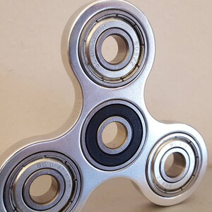 May include: A silver fidget spinner with three ball bearings. The bearings have the text "BC Precision" and "Z208" inscribed on them.