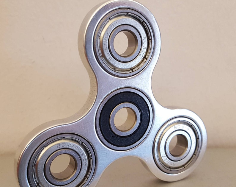May include: A silver fidget spinner with three ball bearings. The bearings have the text "BC Precision" and "Z208" inscribed on them.