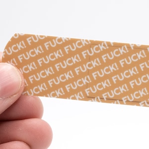 May include: A tan bandage with the word "FUCK!" repeated in white. The bandage is held by a person's hand against a white background. The text is in a bold, sans-serif font.