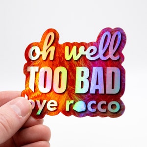 May include: A holographic sticker with the text "oh well TOO BAD bye Rocco" in a rainbow color scheme.