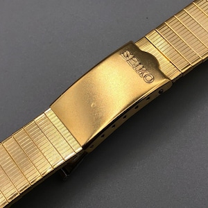 May include: Gold-toned Seiko watch band with a textured, expandable design. The clasp features the Seiko logo. The band is made of small, rectangular links, creating a flexible and comfortable fit. The watch band is a classic accessory.