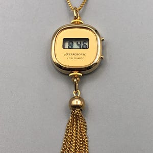 May include: A gold pendant necklace with a digital watch face showing "8:46". The watch is inscribed with "ASTROSONIC LCD QUARTZ" and has a gold tassel. The chain is also gold.