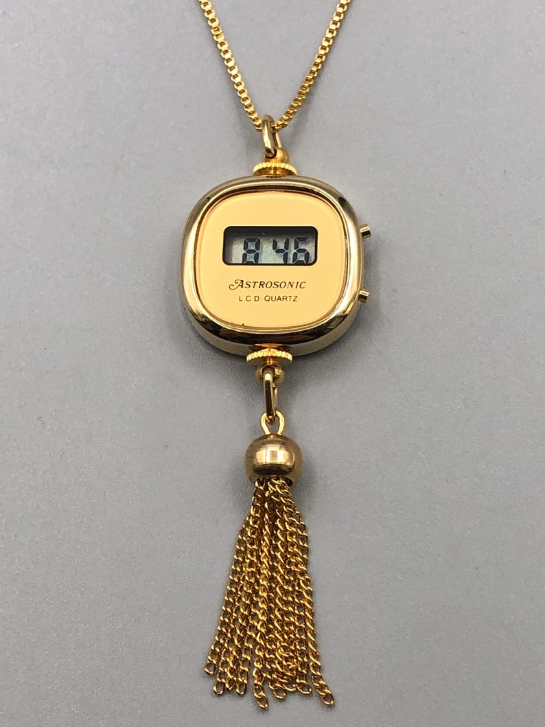 May include: A gold pendant necklace with a digital watch face showing "8:46". The watch is inscribed with "ASTROSONIC LCD QUARTZ" and has a gold tassel. The chain is also gold.