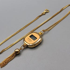 May include: Gold-coloured necklace featuring a digital watch pendant. The watch shows the time and is labelled "Astronic LCD Quartz". A gold chain and tassel complete the design.