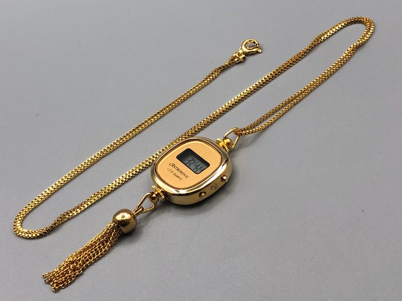 May include: Gold-coloured necklace featuring a digital watch pendant. The watch shows the time and is labelled "Astronic LCD Quartz". A gold chain and tassel complete the design.