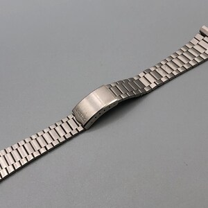 May include: A silver-toned metal watch band with a folding clasp. The band is made of small, rectangular links. The clasp is marked with the word "ORIENT". The watch band is designed to be attached to a watch case.