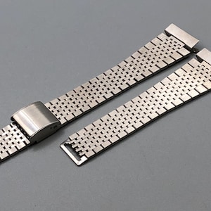 May include: A stainless steel watch band with a mesh design. The band is composed of small, rectangular links and features a clasp. The band is shown in two separate pieces, with one piece connected to the clasp.