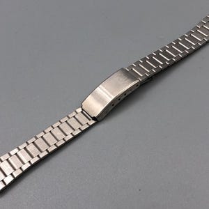 May include: A silver-toned watch band with a rectangular link design. The band features a folding clasp and is made of a shiny, metallic material. The watch band is shown against a gray background.