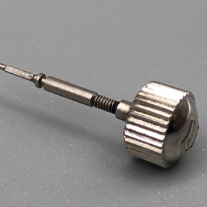 May include: A silver-toned watch crown with a ridged edge and a small, detailed tip. The crown has a threaded shaft and a small, cylindrical end. The watch part is designed for precision and durability.