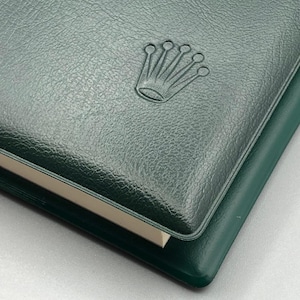 May include: A dark green leather-bound notebook with a debossed crown logo. The notebook has a textured surface and a contrasting lighter-colored edge. The cover is slightly overlapping another dark green surface.
