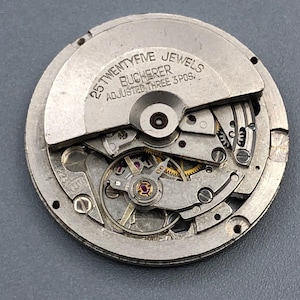 K&ouml;nnte beinhalten: Nahaufnahme eines Bucherer-Uhrwerks mit sichtbaren Zahnr&auml;dern, Juwelen und komplizierten mechanischen Komponenten. Das silberfarbene Metallwerk tr&auml;gt den Text "25 TWENTYFIVE JEWELS BUCHERER ADJUSTED THREE 3 POS."