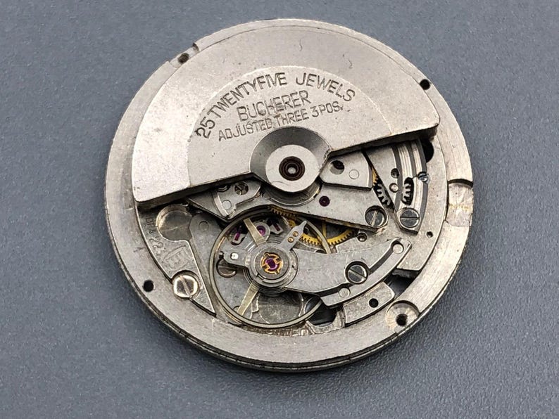 K&ouml;nnte beinhalten: Nahaufnahme eines Bucherer-Uhrwerks mit sichtbaren Zahnr&auml;dern, Juwelen und komplizierten mechanischen Komponenten. Das silberfarbene Metallwerk tr&auml;gt den Text "25 TWENTYFIVE JEWELS BUCHERER ADJUSTED THREE 3 POS."