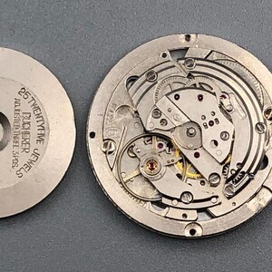 K&ouml;nnte beinhalten: Zerlegtes Uhrwerk mit filigranen Zahnr&auml;dern und Komponenten. Der silberfarbene Mechanismus zeigt Schrauben, Federn und eine Unruh. Ein separates silberfarbenes Teil tr&auml;gt die Aufschrift "25 TWENTY-FIVE JEWELS BUCHERER ADJUSTED THREE 3 POS."