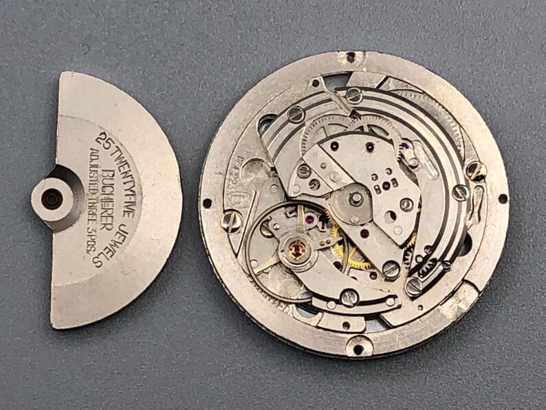K&ouml;nnte beinhalten: Zerlegtes Uhrwerk mit filigranen Zahnr&auml;dern und Komponenten. Der silberfarbene Mechanismus zeigt Schrauben, Federn und eine Unruh. Ein separates silberfarbenes Teil tr&auml;gt die Aufschrift "25 TWENTY-FIVE JEWELS BUCHERER ADJUSTED THREE 3 POS."